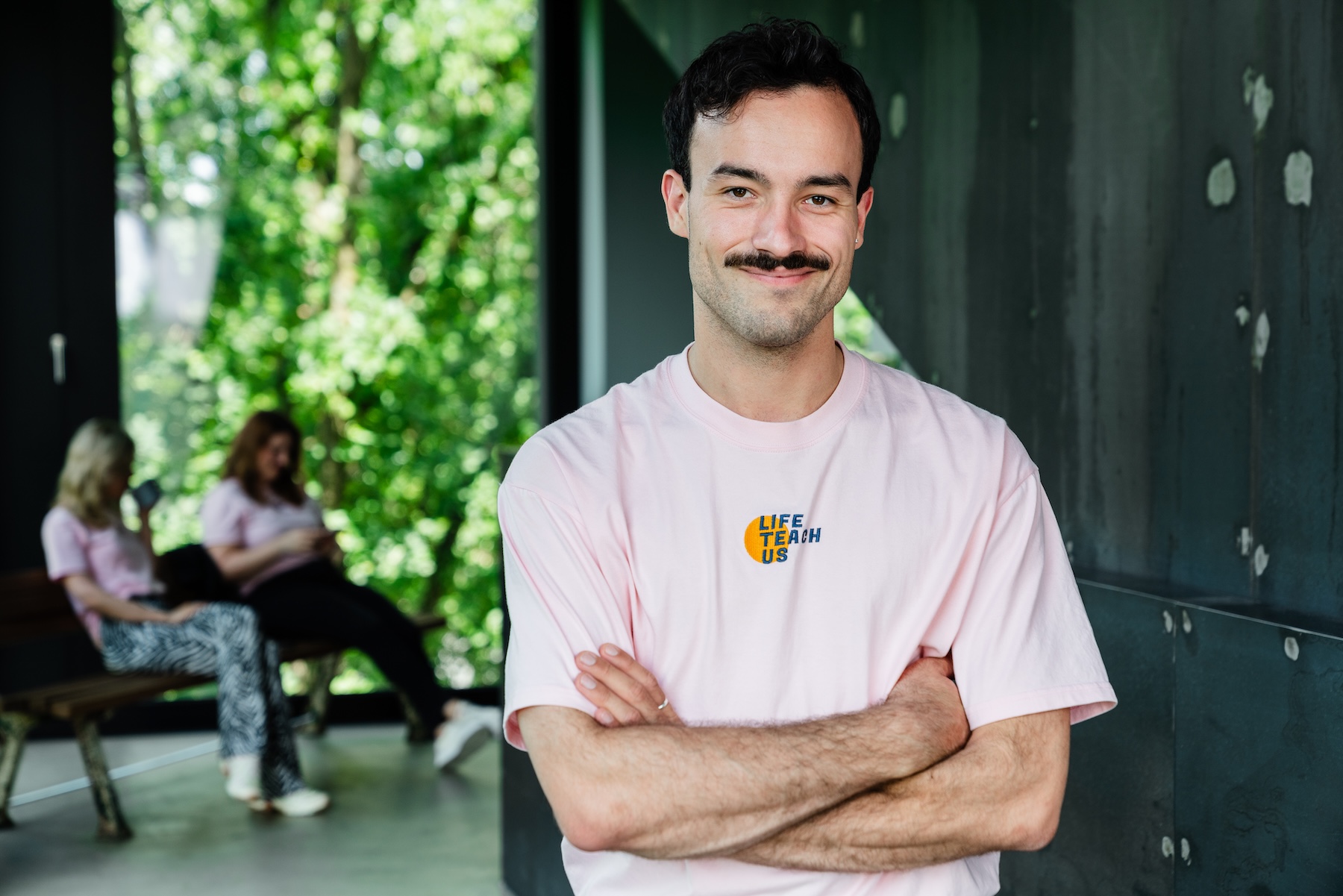 Ludwig Thiede, Geschäftsführer der LifeTeachUs gGmbH, steht in einem rosa Shirt in einem Raum und hat die Arme vor dem Oberkörper verschränkt. Links im Hintergrund sitzen zwei weiblichen Personen auf einer Bank.