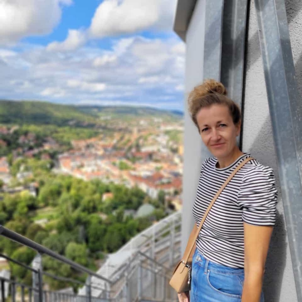 Portrait von Claudia, Teilnehmerin des Online-Programms Mit Klarheit in deine berufliche Zukunft. Sie trägt ein scharz-weißes gestreiftes Shirt, eine Jeans und hat eine hellbraune Umhängetasche. Sie steht auf einer Plattform und Hunter ihr ist ein Stadtpanorama zu sehen.