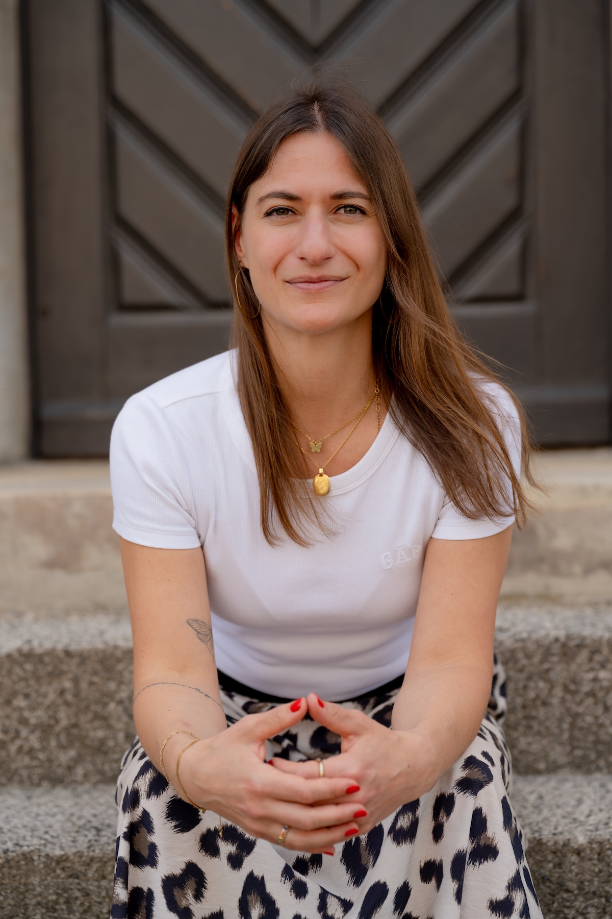 Giulia Barile, DGSv-zertifizierte systemische Supervisorin und Coach und Mentorin für Frauen in der beruflichen Neuorientierung, trägt ein weißes Shirt, einen Printrock, sitzt auf einer Steintreppe vor einer braunen Holztür