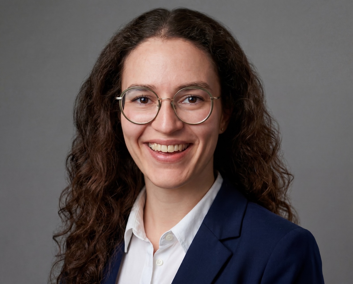 Portrait von Elena Haas. Sie trägt eine weiße Bluse und eine blauen Blazer. Sie trägt eine Brille, die lockigen Haare offen und lächelt direkt in die Kamera.